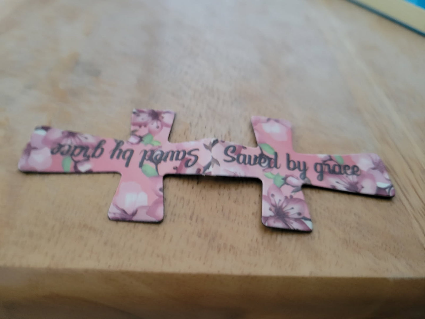 Pink floral cross-shaped magnetic bookmark with text on a wooden surface.