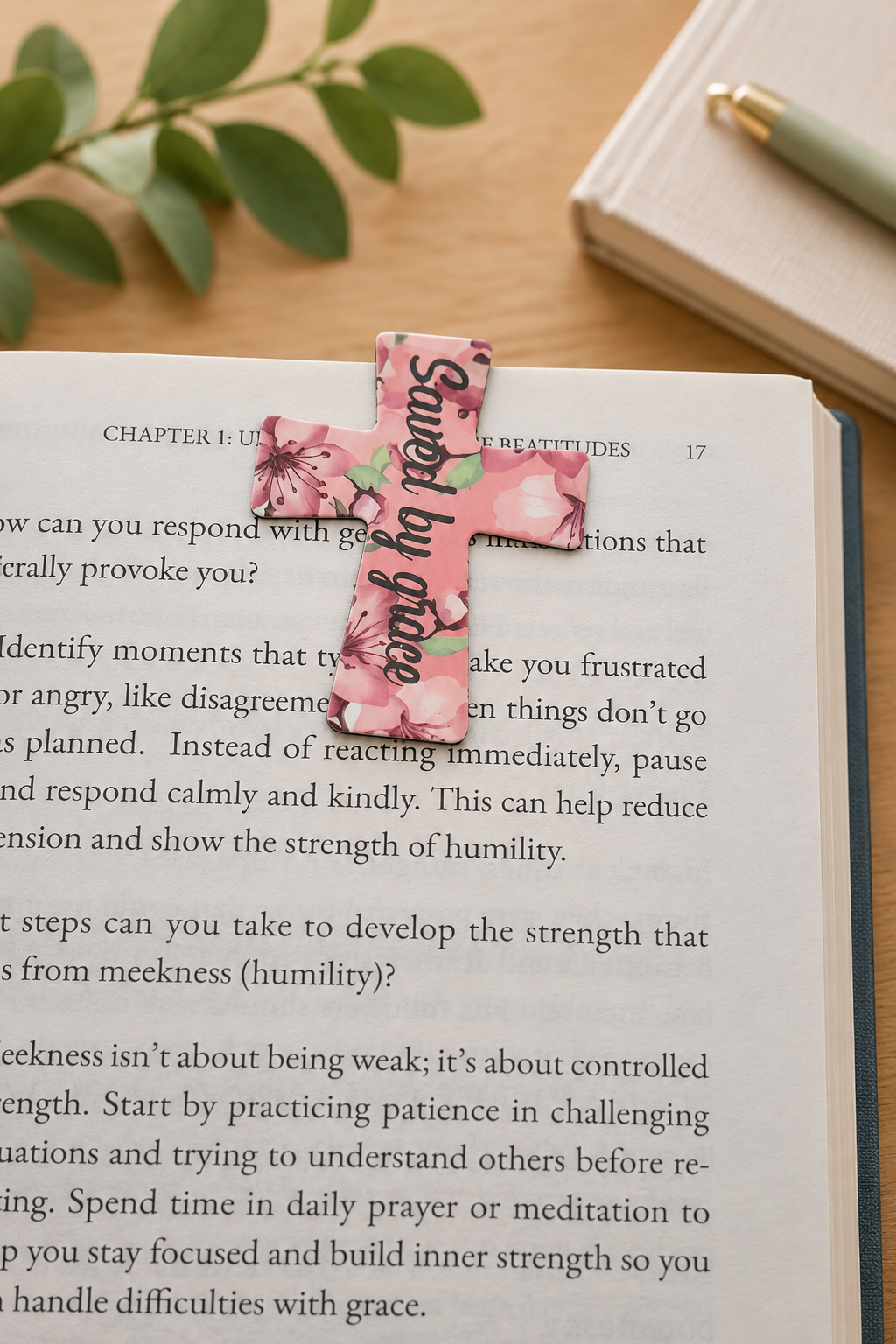 Pink floral “Saved by Grace” cross magnetic bookmark marking a page in an open book on a light wood table.