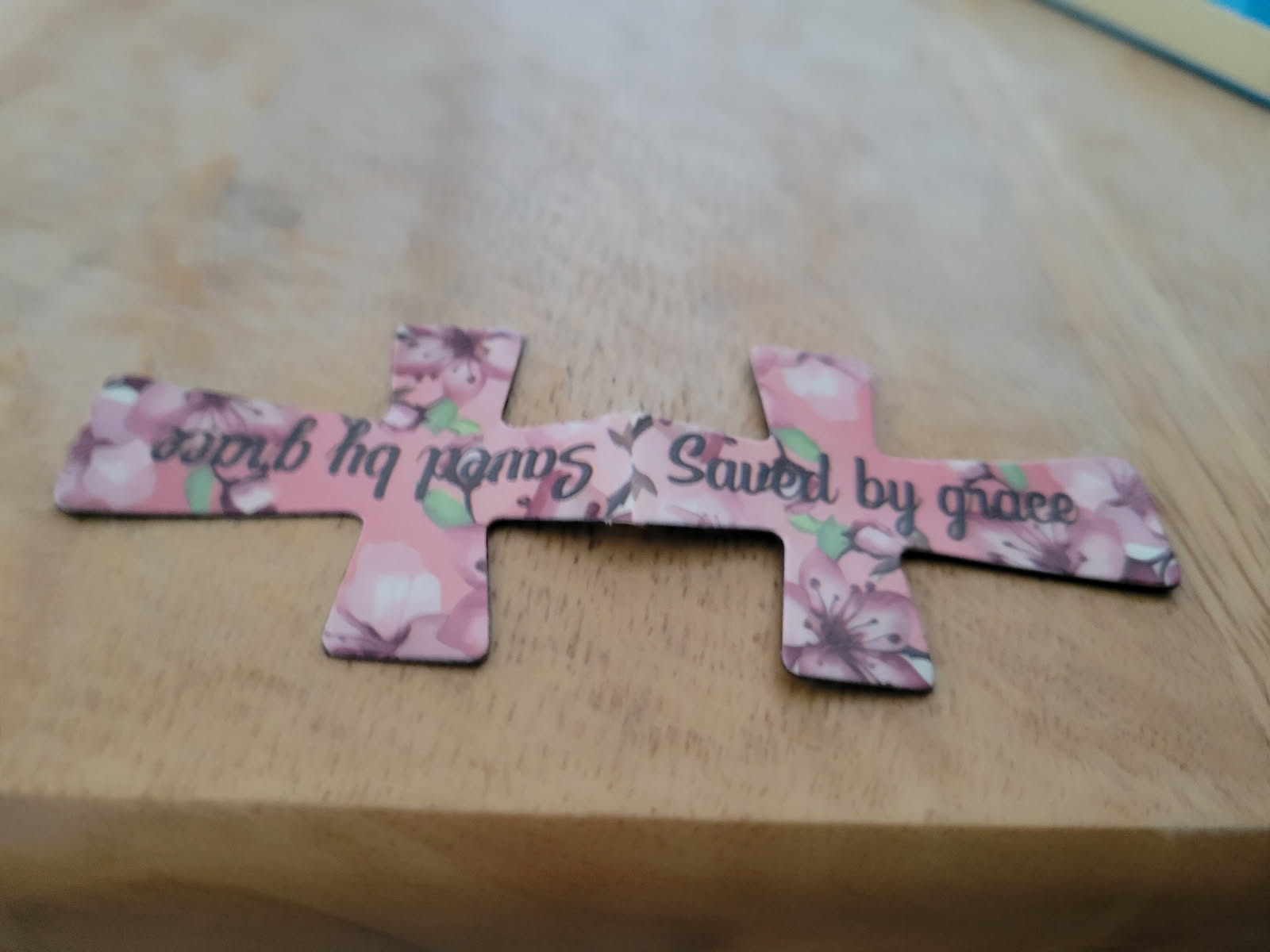 Pink floral cross-shaped magnetic bookmark with text on a wooden surface.